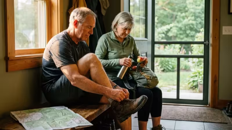 An active senior couple in their 60s prepares for a hike in a sunlit mudroom, lacing up boots and packing a bag.