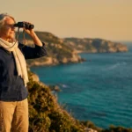 An active senior woman looks through binoculars from a coastal cliff over the Mediterranean sea during golden hour.