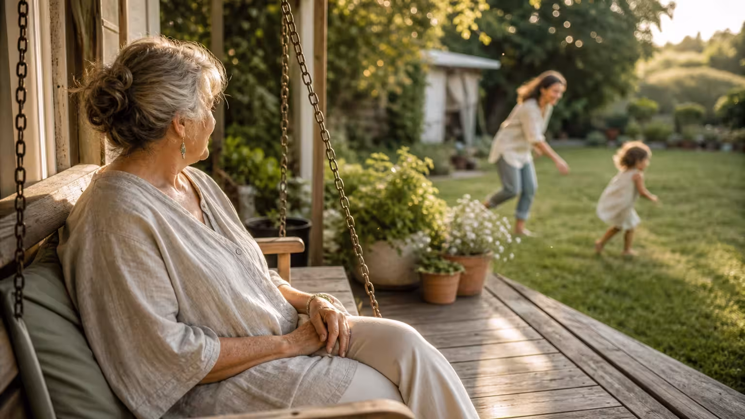 A grandmother sits peacefully on a porch swing watching her daughter and grandchild play in the yard during golden hour.